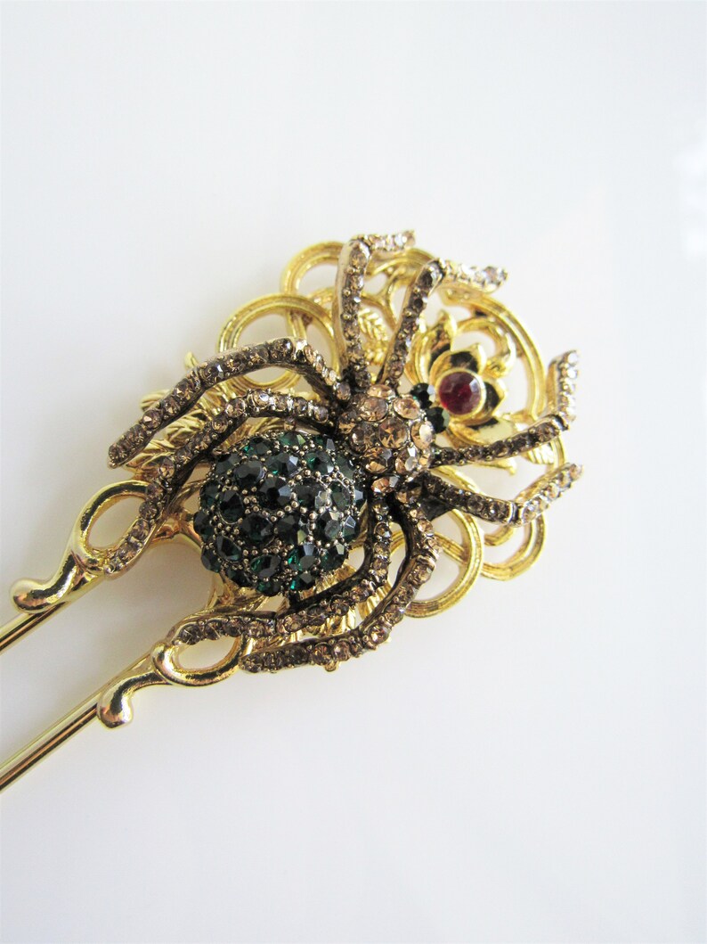 Puede incluir: Varilla para el pelo de tono dorado con un dise&ntilde;o de ara&ntilde;a adornada con gemas. La ara&ntilde;a tiene un cuerpo de gema verde y una gema roja en el centro de su cabeza. La ara&ntilde;a est&aacute; rodeada de un dise&ntilde;o de filigrana decorativo.