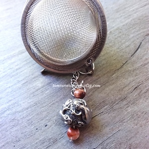 May include: A silver tea infuser with a mesh ball and a chain attached to a silver bead with a brown bead on either side. The bead has a detailed design of two fish.