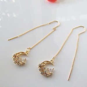 May include: A pair of gold-colored threader earrings. Each earring features a crescent moon design with star and crystal accents. The earrings have a delicate chain that threads through the ear.