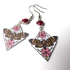 May include: A pair of silver earrings with a red gemstone and a white diamond-shaped pendant featuring a black and brown moth with pink flowers.