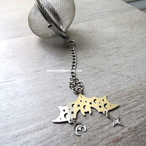 May include: Silver tea infuser with a chain and a bat-shaped charm with stars and moons. The charm is attached to the chain with a small silver ring.