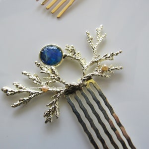 May include: A gold hair comb with a tree branch design. The comb has a blue gemstone in the center and small pearl-like accents on the branches.