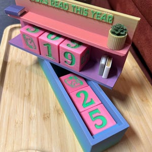 May include: A pink and green wooden bookshelf-style display with the text "BOOKS READ THIS YEAR". It features pink blocks with green numbers, a small succulent, and miniature books. A blue tray holds additional blocks.