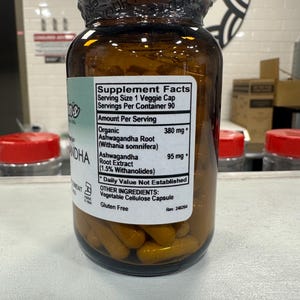 May include: A brown glass bottle with a white label that reads "Supplement Facts" and lists the ingredients "Organic Ashwagandha Root (Withania somnifera) 380 mg*" and "Ashwagandha Root Extract (1.5% Withanolides) 95 mg*" along with other ingredients and a "Gluten Free" label. The bottle is filled with yellow capsules.
