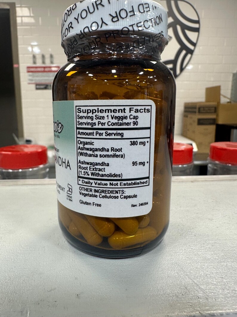 May include: A brown glass bottle with a white label that reads "Supplement Facts" and lists the ingredients "Organic Ashwagandha Root (Withania somnifera) 380 mg*" and "Ashwagandha Root Extract (1.5% Withanolides) 95 mg*" along with other ingredients and a "Gluten Free" label. The bottle is filled with yellow capsules.
