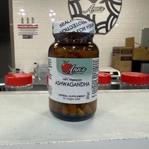 May include: A brown bottle with a white label that says "Ann's Since 1984" and "100% Vegetarian Ashwagandha Herbal Supplement 90 Veggie Caps". The bottle is filled with yellow capsules and has a red apple logo on the label.