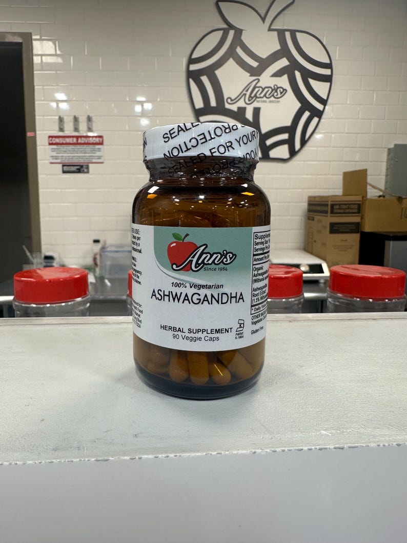 May include: A brown bottle with a white label that says "Ann's Since 1984" and "100% Vegetarian Ashwagandha Herbal Supplement 90 Veggie Caps". The bottle is filled with yellow capsules and has a red apple logo on the label.