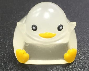 May include: A white, translucent penguin figurine with yellow feet and a yellow beak. The penguin is sitting on a white base.