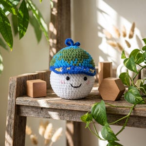 Puede incluir: Una esfera blanca tejida con una cara sonriente, que lleva un sombrero tejido azul y verde. El sombrero tiene un borde azul con detalles amarillos. La esfera tiene mejillas rosadas y ojos negros. El artículo se exhibe en un estante de madera rústica.
