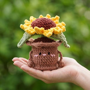 Puede incluir: Un girasol de ganchillo en una maceta marrón con una cara sonriente. El girasol tiene pétalos amarillos y un centro marrón. La maceta tiene brazos y piernas. El artículo se sostiene en una mano sobre un fondo verde borroso.