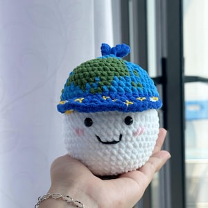 Op de afbeelding: Een gehaakte aarde-knuffel met een lachend gezicht, met een blauw en groene hoed. De hoed is ontworpen om op de aarde te lijken, met blauwe oceanen en groene continenten. De knuffel is wit en wordt in een hand gehouden.