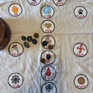 May include: A cream-coloured cloth with hand-painted circular designs featuring symbols and runes in black, red, blue, and yellow. Several small, dark stones and wooden objects are scattered across the cloth. The theme suggests divination or spiritual practices.