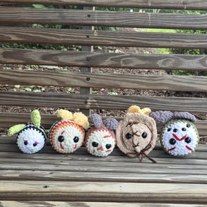 May include: Five round, crocheted plush toys featuring various designs including a black and white striped pattern, a white and orange creature, a white and red patterned figure, a tan stitched character, and a green and white hockey mask design.