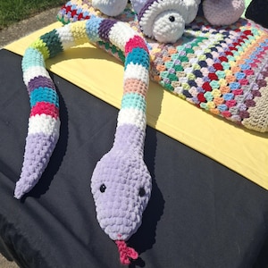 May include: A large, soft, crocheted snake in shades of lavender, white, and rainbow colors. The snake has black eyes and a pink tongue. Other crocheted items, including mushrooms and a blanket, are also visible.