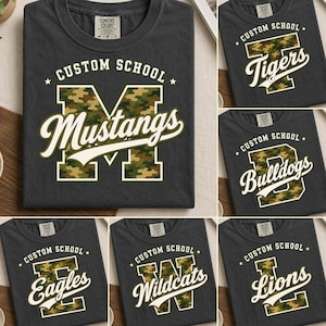 May include: A dark gray t-shirt featuring a large camouflage-patterned block letter with the text "CUSTOM SCHOOL" above and a team name like "Mustangs," "Tigers," "Bulldogs," "Eagles," "Wildcats," or "Lions" in white script across the letter.