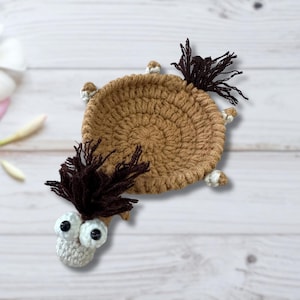 May include: A handmade crochet coaster in the shape of a turtle. The coaster is primarily brown with dark brown fringe and white and brown accents. The turtle has large, googly eyes.