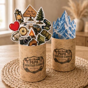 May include: Two cylindrical containers with a rustic design, filled with camping-themed cutouts and a mountain-shaped card. The cutouts include a lantern, compass, camera, and a sign that reads "Für den besten Papa". The containers have text that reads "Mit PAPA ist jedes Abenteuer schöner".