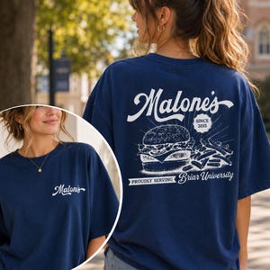 Op de afbeelding: Marineblauw t-shirt met een witte afbeelding van een hamburger en friet, en de tekst "Malone's Since 2015". Het shirt heeft ook de tekst "Proudly Serving Briar University". Het shirt wordt door een persoon gedragen.
