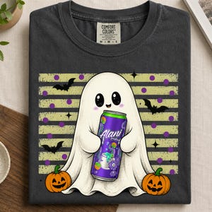 Op de afbeelding: Een antracietgrijs t-shirt met een afbeelding van een witte geest die een paars Alani Nu energiedrankblikje vasthoudt. Het ontwerp bevat twee oranje pompoenen, zwarte vleermuizen en een achtergrond van horizontale beige strepen met paarse stippen.