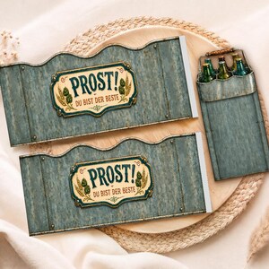 May include: Two blue-grey beer bottle carriers with the text "PROST! DU BIST DER BESTE" in a decorative frame. A matching carrier holds several green bottles. The items are on a wooden tray with a woven border.