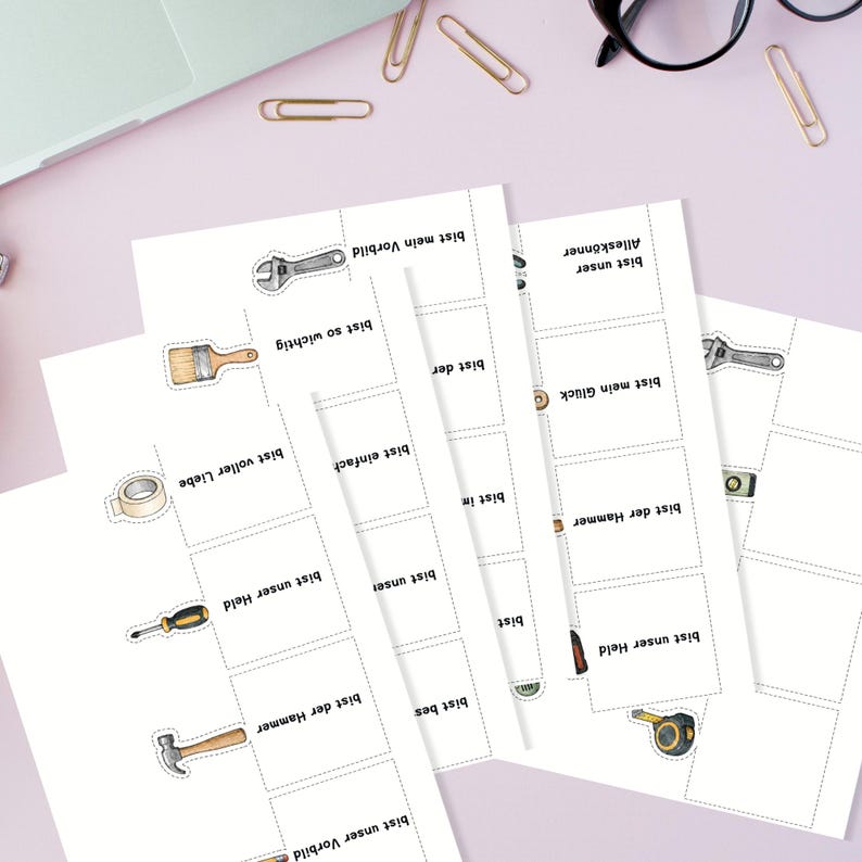 May include: A collection of white cards with illustrations of tools and German text. The tools include a wrench, paintbrush, tape, screwdriver, hammer, and measuring tape. The cards are arranged on a pink surface with office supplies.