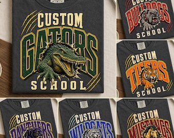 May include: A charcoal gray t-shirt featuring a graphic print of a green alligator with the text CUSTOM GATORS SCHOOL. The design includes claw marks and bold lettering. Other variations show a bulldog, tiger, panther, wildcat, or mustang.