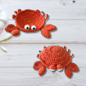 May include: Two handmade, orange crochet crab coasters. Each coaster features a round body, large claws, and small eyes. The coasters are designed to resemble crabs.