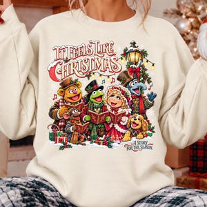 Könnte beinhalten: Ein cremefarbenes Sweatshirt mit Rundhalsausschnitt und einer bunten Grafik von Muppet-Figuren, die Weihnachtslieder singen. Der Text lautet It Feels Like Christmas und A Story For The Season. Das Design zeigt Fozzie Bär, Kermit, Miss Piggy, Gonzo und einen kleinen Hund.