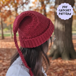 以下が含まれることがあります： 長い三つ編みのテールが付いた、バーガンディ色の手編みの帽子。 帽子にはリブ編みのつばと丸みを帯びたクラウンがあります。 テキスト「PDF CROCHET PATTERN」は薄紫色の円の中にあります。 背景は、木と落ち葉のあるぼやけた秋のシーンです。