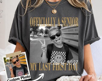May include: A charcoal gray short-sleeve t-shirt featuring a large rectangular graphic with the text "OFFICIALLY A SENIOR" and "MY LAST FIRST DAY" in gold. The graphic shows a black and white photo of a student wearing sunglasses and a smiley face shirt.