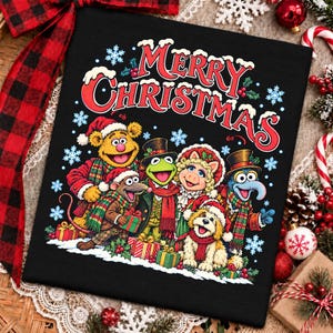 Può includere: Una t-shirt nera con una grafica colorata dei personaggi dei Muppet, tra cui Fozzie Bear, Kermit la rana, Miss Piggy e Gonzo che indossano abiti invernali, con la scritta Merry Christmas in lettere rosse sopra e dei fiocchi di neve.