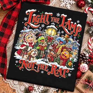 Può includere: Una t-shirt nera con una grafica festiva dei Muppets che ritrae Miss Piggy, Kermit, Gonzo, Fozzie Bear e un cane vicino a un lampione. Il testo "LIGHT THE LAMP NOT THE RAT" è scritto in lettere rosse e bianche circondate da fiocchi di neve.