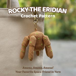 May include: A crocheted keychain featuring a brown, space-themed creature with green accents. The keychain is attached to a gold-colored clasp. Text on the image reads "ROCKY THE ERIDIAN Crochet Pattern" and "Amaze, Amaze, Amaze! Your Favorite Space Friend in Yarn."