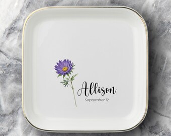 May include: A square white ceramic trinket dish with a gold-toned rim. The center features a purple aster flower illustration, the name Allison in black script, and the text September 12 printed in a simple black sans-serif font below the name.