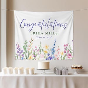 May include: A white banner with the word "Congratulations" in purple script, along with the name "ERIKA MILLS" and "Class of 2026". The banner has a watercolor floral border with pink, purple, yellow, and blue flowers. A cake and cupcakes are on a table.