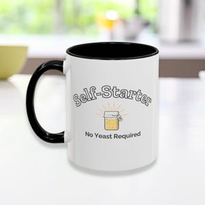 May include: White ceramic mug with a black interior and handle. The mug features the text "self-Starter" above an illustration of a jar with the text "No Yeast Required". The mug is on a white surface.