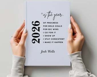 May include: A white journal with the year 2026 printed in black. The journal's cover features the text "*is the year..." followed by motivational phrases. The author's name, Josh Wells, is printed at the bottom. The journal is held by two hands.