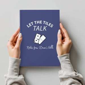 May include: A navy blue notebook with the text "LET THE TILES TALK" and "Notes from Dave's table" in white. The cover features a domino graphic. The notebook is held by a person.