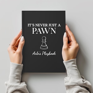 May include: A black notebook with the text "IT'S NEVER JUST A PAWN" and "Arlo's Playbook" in white. A white chess pawn graphic is centered below the text. The notebook is held by two hands.