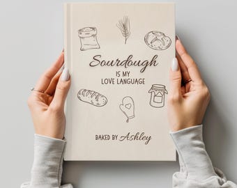 May include: A cream-colored book with the text "Sourdough is my Love Language" and illustrations of bread, a bag of flour, and a jar of jam. The book is held by two hands.