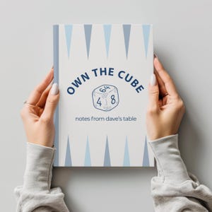 May include: A notebook with a white cover featuring the text "OWN THE CUBE" and a dice graphic. Below the graphic is the text "notes from dave's table". The cover has a light blue and white geometric design. The notebook is held by two hands.