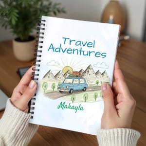 May include: A spiral-bound notebook with a travel-themed illustration. The cover features a blue car driving on a road, mountains, a sun, and the text "Travel Adventures" and "Makayla" in teal and green.