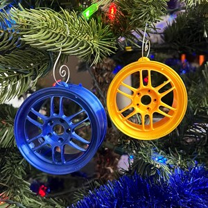 Puede incluir: Dos adornos de rueda de coche, uno azul y otro amarillo, cuelgan de un árbol de Navidad. Los adornos son redondos con un diseño detallado de radios. El árbol está decorado con luces y oropel azul.