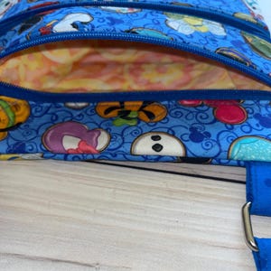 Puede incluir: Una bolsa de tela azul con cierre de cremallera. La bolsa presenta un colorido estampado de personajes de dibujos animados. El forro interior es un estampado floral melocot&oacute;n y amarillo. Una correa azul es visible en el lateral.