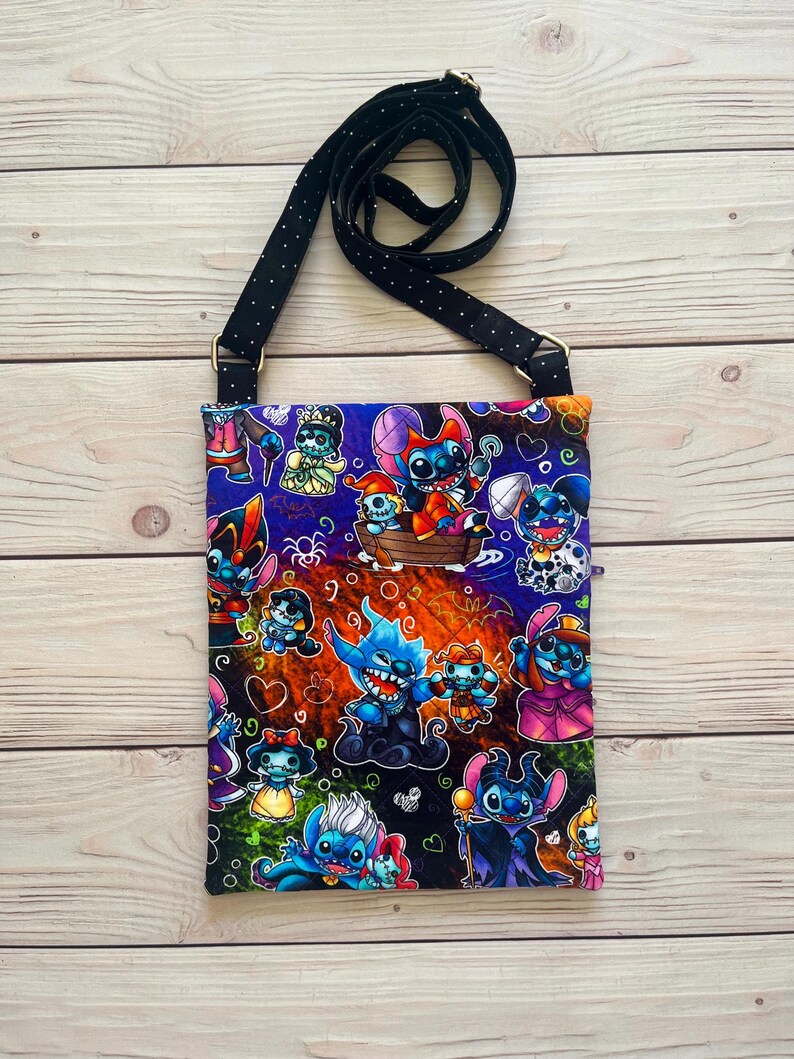 Puede incluir: Un bolso bandolera rectangular con una correa negra con lunares blancos. La tela del bolso est&aacute; estampada con personajes de dibujos animados coloridos sobre un fondo oscuro. El bolso tiene herrajes met&aacute;licos dorados.