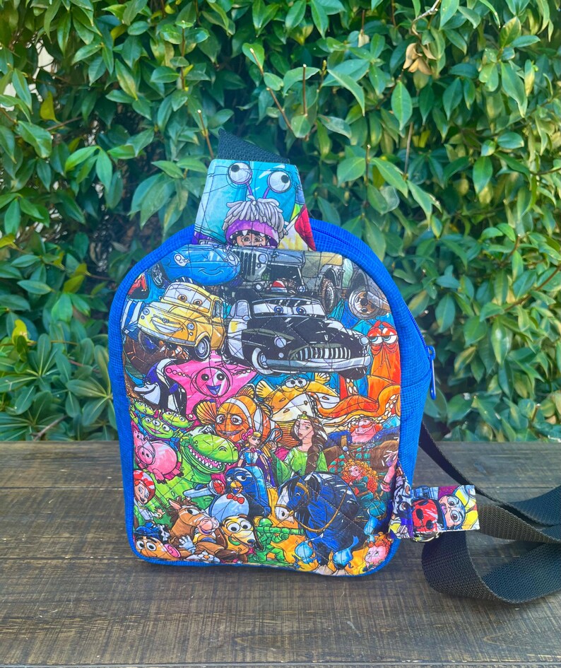 May include: A small blue backpack with a colorful print of various Disney characters, including Cars, Toy Story, and The Little Mermaid. The backpack has a single adjustable strap.