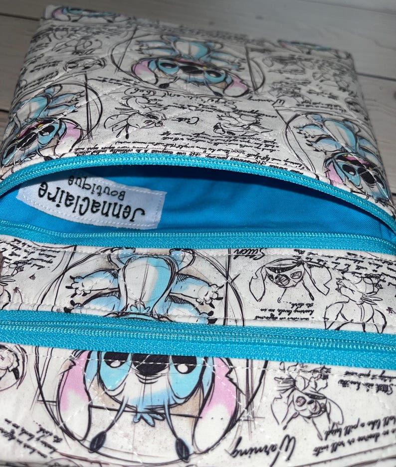 May include: A quilted pouch featuring a white background with a repeating blue and pink character print and black cursive text. The pouch has a bright blue interior lining, a matching blue zip, and a white label reading jennaclaire boutique.