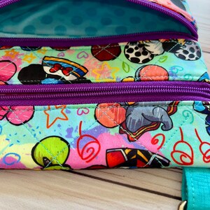 K&ouml;nnte beinhalten: Nahaufnahme einer bunten, gesteppten Umh&auml;ngetasche mit einem lebendigen Cartoon-Charakter-Print. Die Tasche verf&uuml;gt &uuml;ber mehrere Rei&szlig;verschlussf&auml;cher mit lila Rei&szlig;verschl&uuml;ssen und einen t&uuml;rkisfarbenen Riemen. Das Innenfutter ist hellblau mit Tupfen.