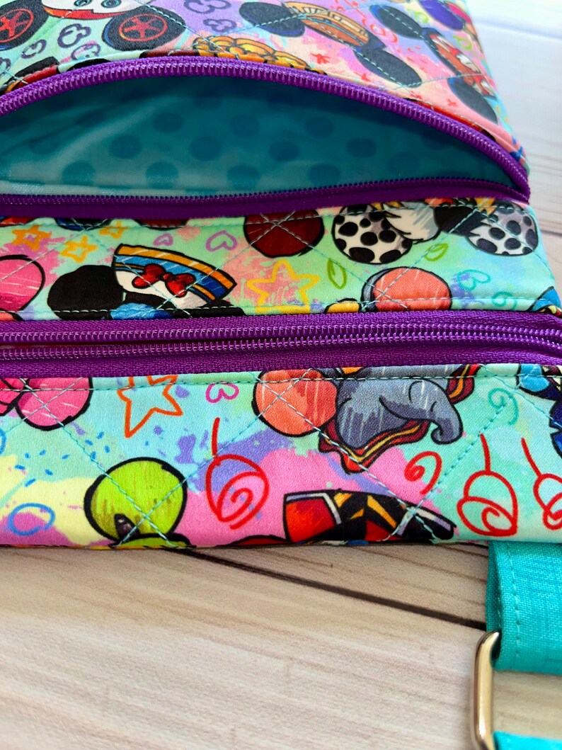 K&ouml;nnte beinhalten: Nahaufnahme einer bunten, gesteppten Umh&auml;ngetasche mit einem lebendigen Cartoon-Charakter-Print. Die Tasche verf&uuml;gt &uuml;ber mehrere Rei&szlig;verschlussf&auml;cher mit lila Rei&szlig;verschl&uuml;ssen und einen t&uuml;rkisfarbenen Riemen. Das Innenfutter ist hellblau mit Tupfen.