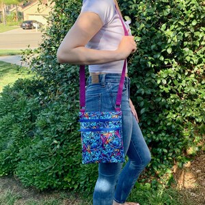 May include: A blue crossbody bag with a colorful pattern featuring cartoon characters. The bag has a purple strap and multiple zippered compartments. The bag is worn over the shoulder.